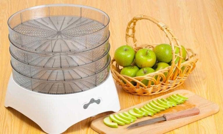 sliced green apple with knife on wooden board next to transparent dehydrator and wooden basket full of green apples