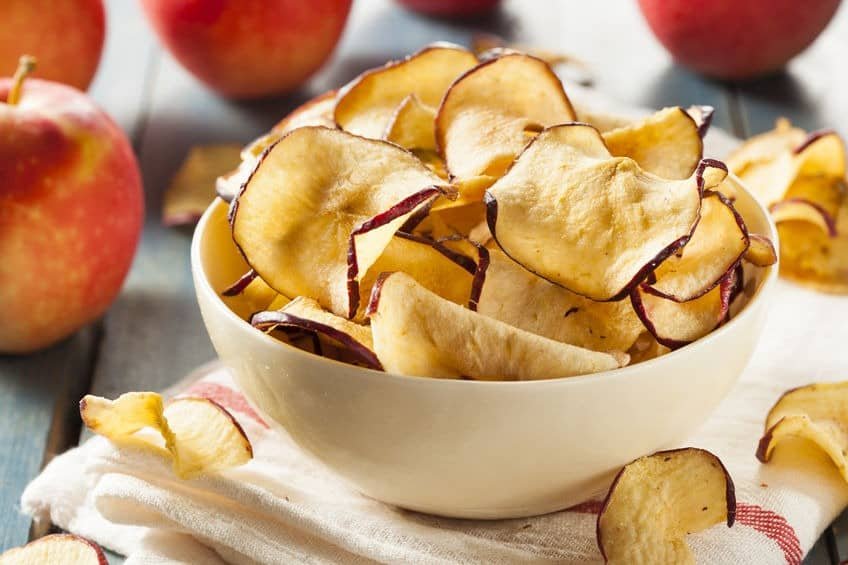 apple chips in a ceramic bowl on a drying cloth with whole red apples next to it on a wooden table