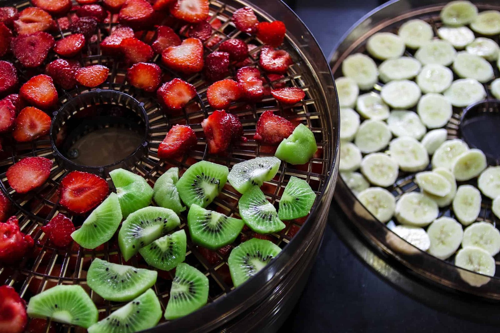 sliced strawberries and kiwis on a dehydrator tray next to it is sliced banana on dehydrator tray on black background