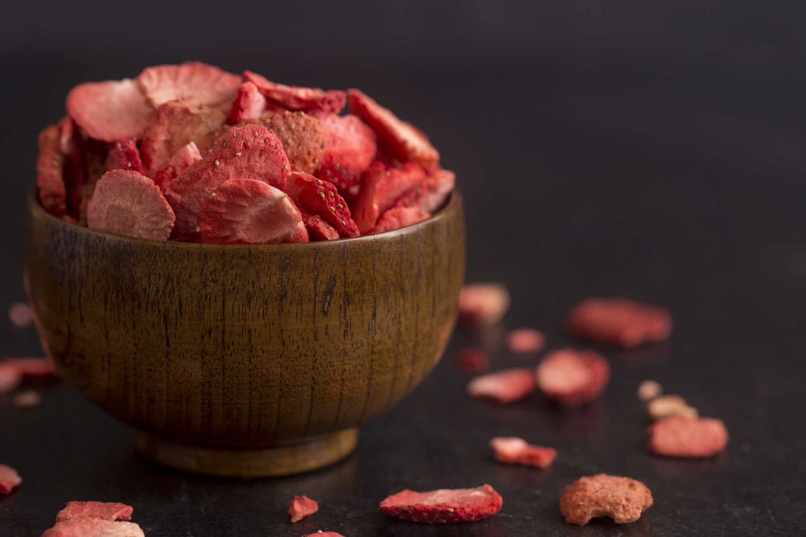 dried strawberries in small wooden bowl and around the bowl on a black background