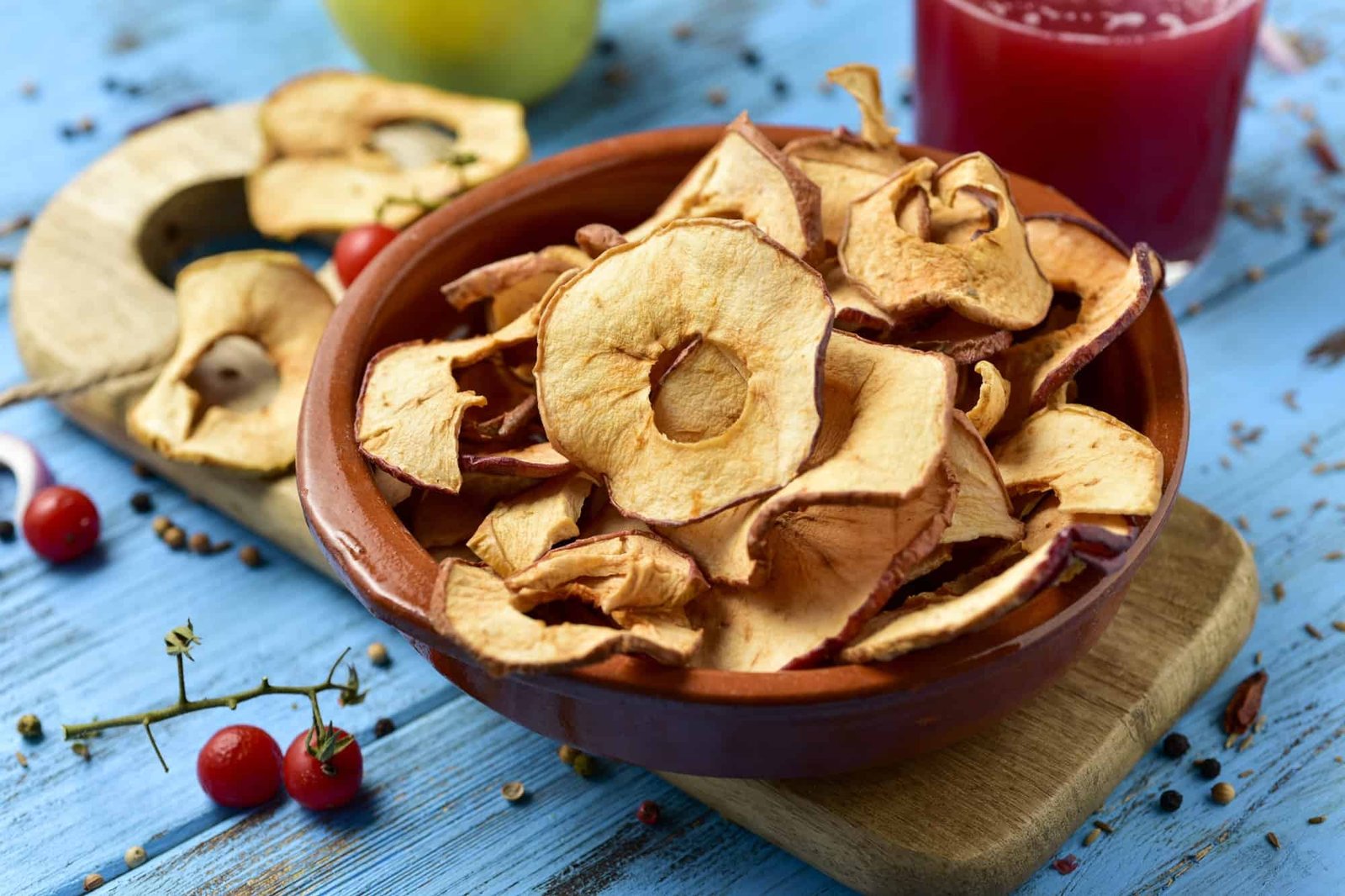 closeup of some slices of dried apple served as appetizer or snack, on a blue rustic wooden table, next to a glass with a red smoothie