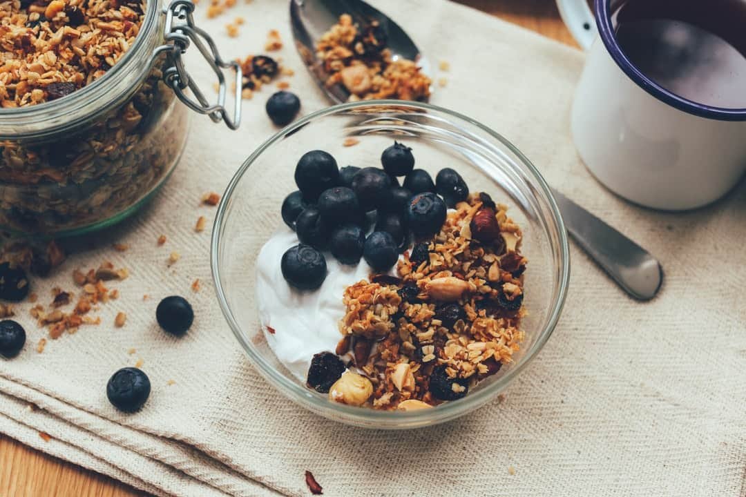 blueberries, granola, yogurt in a glass bowl. glass jar with granola next to bowl and spoon on the drying cloth