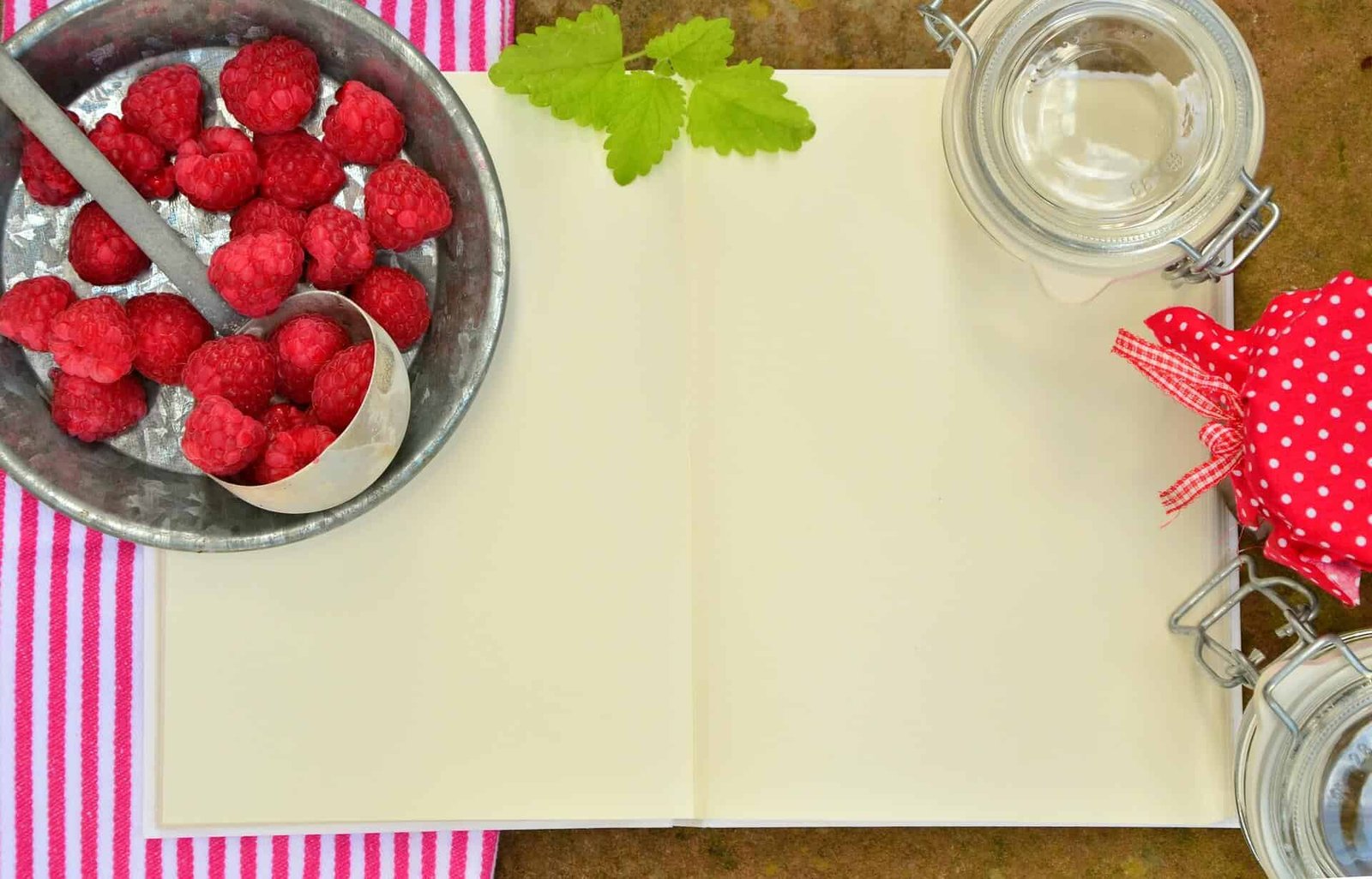 blank open book on top of dish towel, rasberries in a bucket with spoon, glass jar