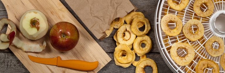 picture of apple skin sliced off on wooden board with orange knife, food dehydrator tray with finished dehydrated apple chips