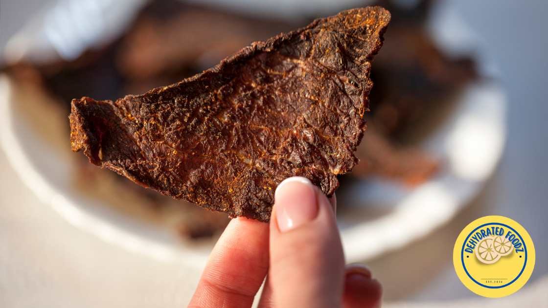 image of a ladies hand holding a piece of beff jerky, in the background is beef jerky in a bowl