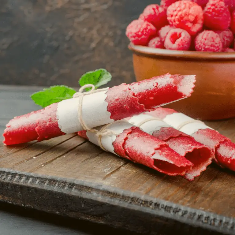 Raspberry fruit leather roll on a wooden table with raspberries in a bowl next to it.