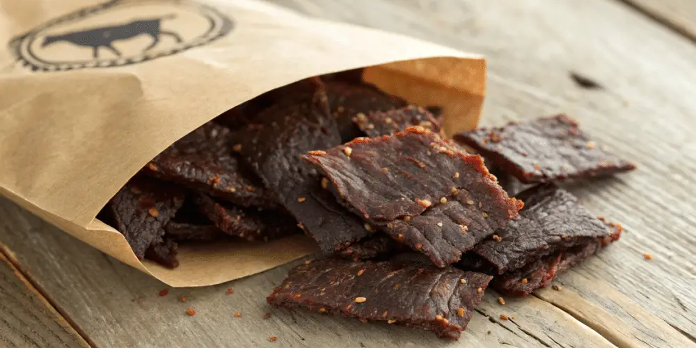 A photo of a bag of dark, spicy beef jerky on a wooden surface. The bag is made of a brown, paper-like material and has a circular logo with a graphic of a silhouette of an animal at the top. The beef jerky is irregularly shaped and has a dark, rich color with seasonings sprinkled on top. The wooden surface provides a rustic and textured backdrop. The image is a close-up, high-resolution photograph with a focus on the beef jerky.