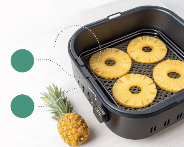 Four pineapple slices on a rack inside a black air fryer with a whole pineapple beside it.