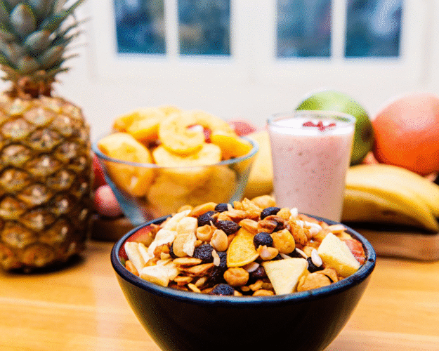 A bowl of granola topped with dried fruits, in front of a smoothie and fresh fruit including bananas, pineapple, and apples.