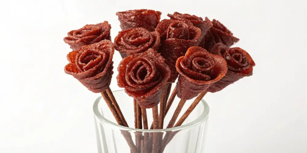 A photo of a creative culinary arrangement featuring several beef jerky snacks shaped into rose-like forms and arranged in a clear glass vase. The brown sticks support the snack roses, giving them a flower-like appearance. The snack roses have a rich, deep brown color and a texture that mimics the layers of a flower's petals. The vase is clear and has a simple, straightforward design. The background is a plain, neutral white. The overall composition is simple and striking.