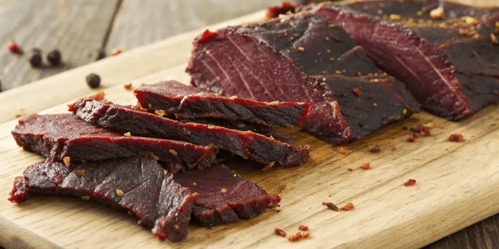 A photo of a close-up of beef jerky on a wooden cutting board. The beef jerky is dark red in color, with visible seasoning and spices scattered across the surface. The cutting board is a natural, light brown wood, with a textured surface. The image is a close-up photograph, capturing the textures and details of the beef jerky and the cutting board. The lighting is warm and natural, highlighting the rich colors of the jerky and the wood.