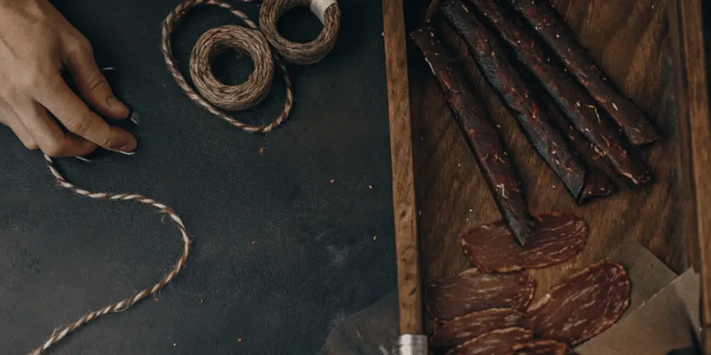 A photograph capturing a close-up view of a culinary scene. A hand is shown in the act of braiding a rope-like structure on a dark, textured surface. The hand is positioned near the braided rope, with a focus on the detailed action of the hand. On the other side of the image, there is a wooden tray with dried meat strips and a rustic, warm brown color. The photograph is a high-resolution, realistic photograph with a focus on textures and colors.