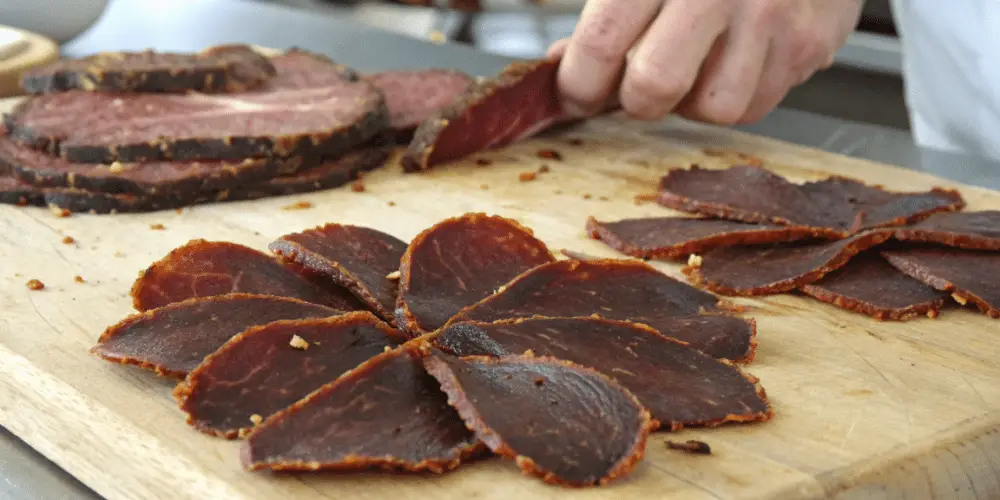 A photograph of a culinary scene with a focus on a wooden cutting board. A hand, with a casual, focused pose, is slicing through a piece of beef jerky. The meat has a rich, dark brown color and a slightly charred appearance. The cutting board is a standard wooden cutting surface with a warm, natural brown hue. The image is a close-up photograph, capturing the textures and details of the meat and the cutting process. The lighting is soft and even.