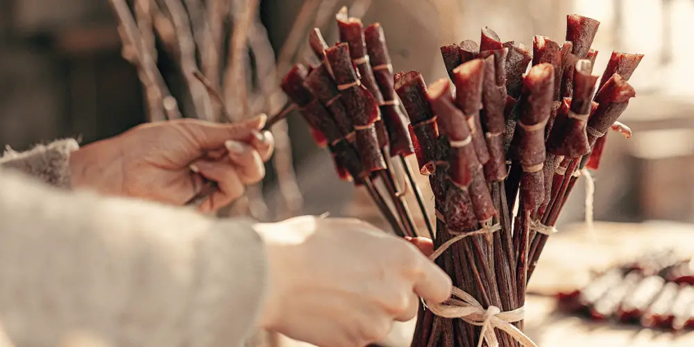 A photograph of a close-up scene of a hand interacting with a bunch of dried, reddish-brown beef jerky, wrapped in twine and displayed in a bunch. The hand, appearing to be that of an adult, is in the process of handling the sausages. The background is softly blurred, featuring muted tones and out-of-focus elements that suggest an indoor setting with a rustic and warm ambiance. The lighting is soft and warm, creating a cozy and inviting atmosphere.