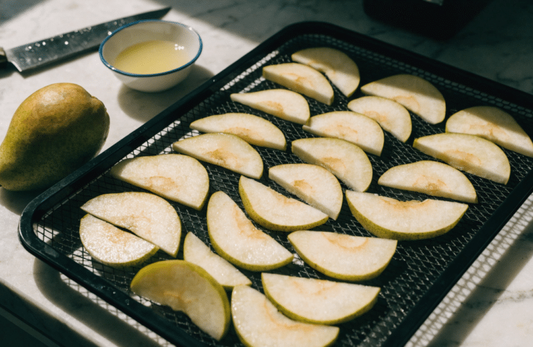 The image shows fresh pears being prepared for dehydration, with sliced pears arranged in a single layer on a dehydrator tray. A sharp knife and a bowl of lemon juice are visible, emphasizing the process of creating delicious, crispy pear chips as a healthy snack.