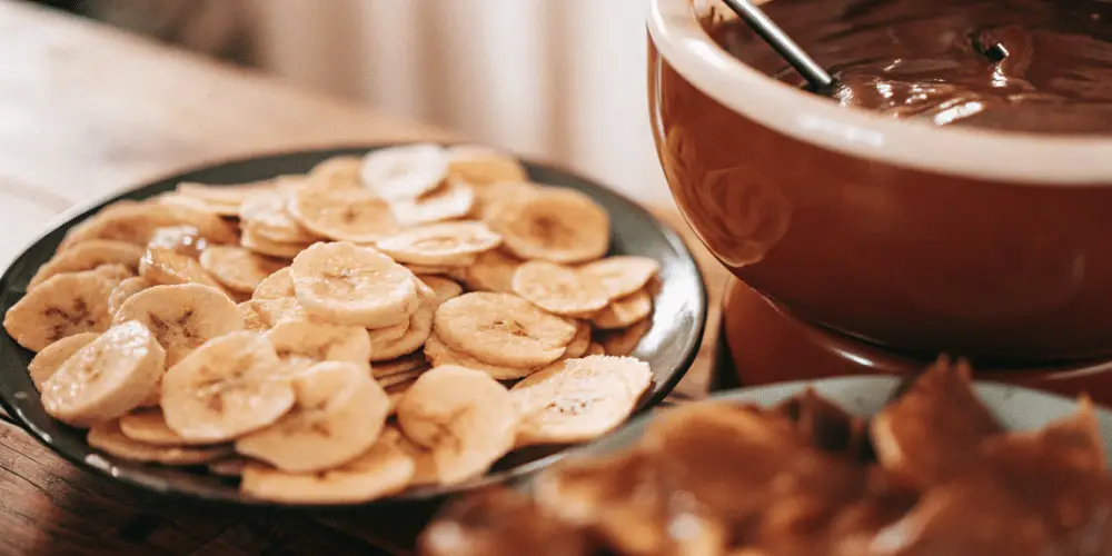 A photo of a table setting with a close-up view of a plate of banana slices and a container with chocolate. The banana slices are arranged on a dark-colored plate, and the chocolate is being served from a brown pot-like container. The table is on a wooden surface, and the background is softly blurred.
