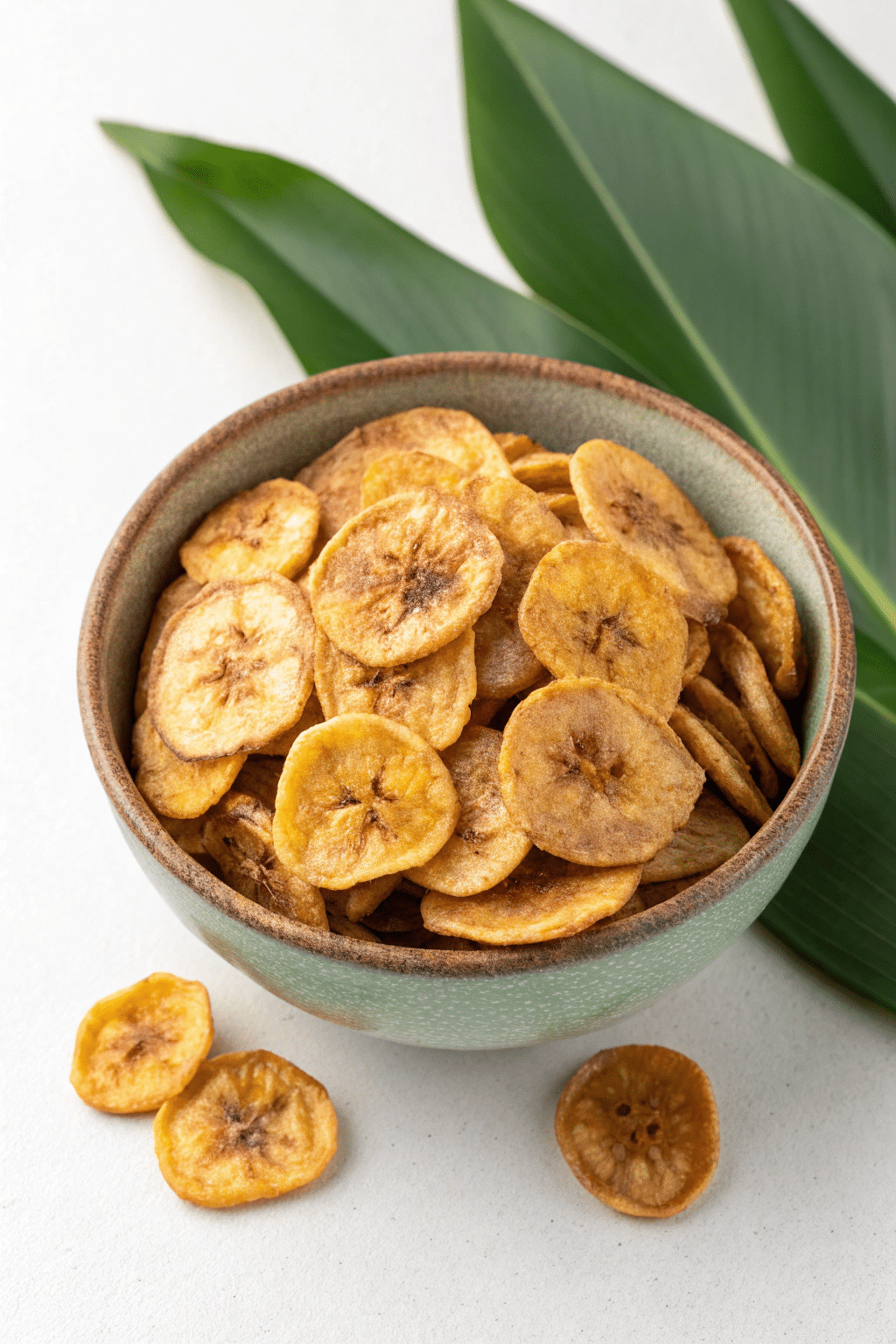 A photo of a bowl filled with banana chips on a white surface, with green leaves in the background. The bowl is made of a textured, matte greenish-gray material and contains numerous golden-brown plantain chips with a slightly charred appearance. Some of the chips have fallen out of the bowl and are scattered around the base of the bowl. The focus is on the plantain chips and the simple, natural elements in the composition.
