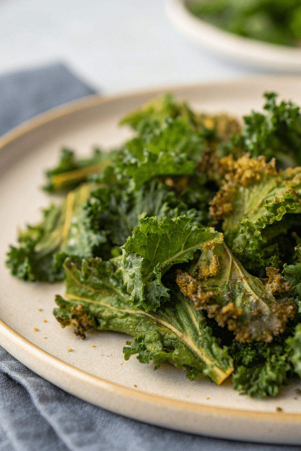 A photo of a culinary scene featuring a plate of cooked kale on a table. The kale is a vibrant green, with some parts showing a slightly browned texture. The plate has a beige rim and is placed on a bluish-gray cloth. The background is softly blurred, focusing the viewer's attention on the plate of kale. The image style is a close-up, high-resolution photograph with a focus on the textures and colors of the kale and the plate.
