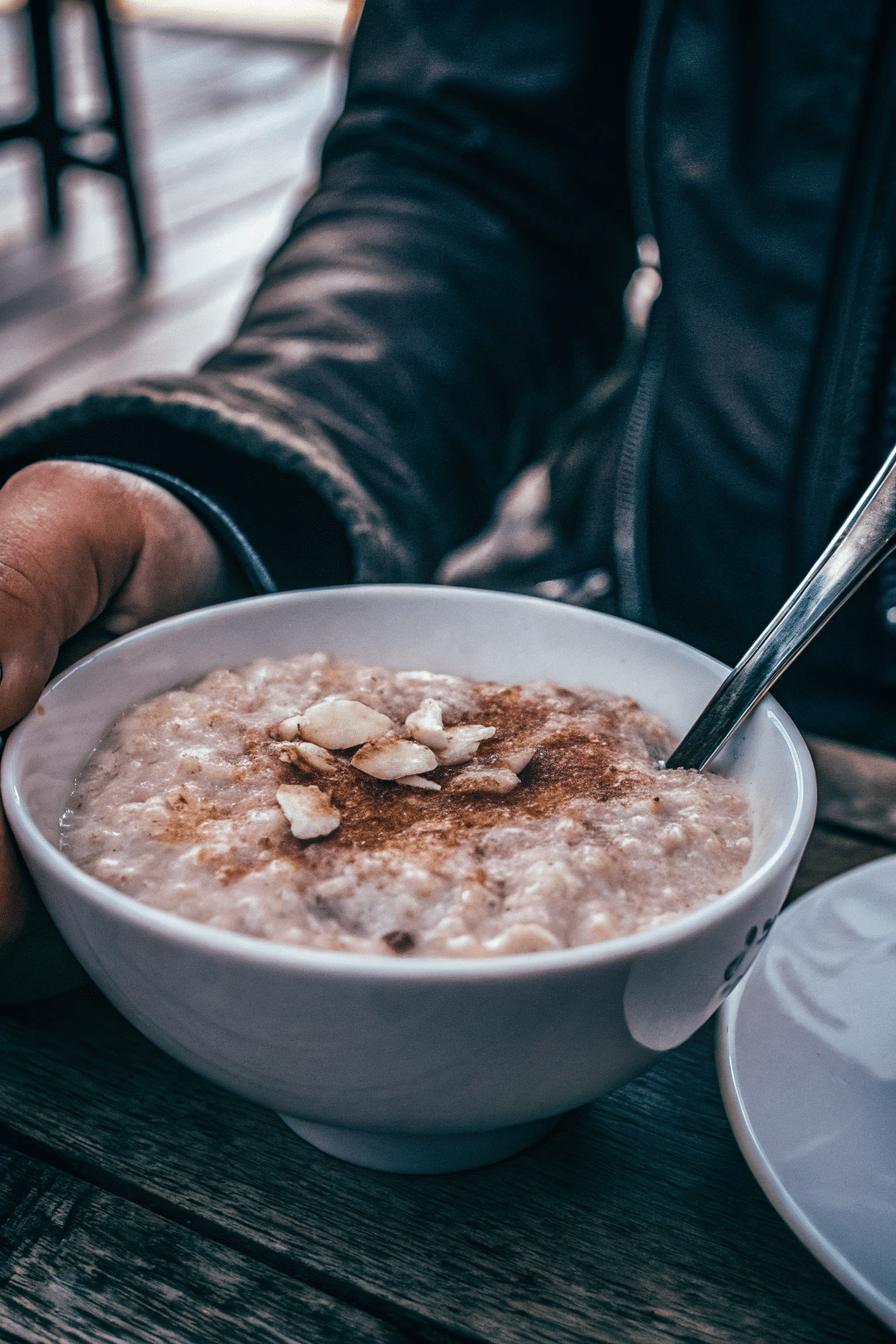A photograph of a close-up scene featuring a person enjoying a meal. The person, partially visible, is wearing a dark, close-fitting leather jacket. The focus is on a white bowl of oatmeal with a warm, brownish topping and several pieces of sliced fruit on top. A spoon is being used to stir the oatmeal. The bowl is placed on a wooden table. The background is softly blurred. The image style is a close-up, high-resolution photograph.