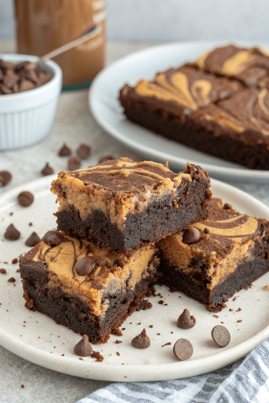 A photo of a close-up view of brownie bars on a white plate. The brownie bars have a rich, chocolatey appearance with a layer of caramel-like topping and scattered chocolate chips. The plate is on a textured, neutral-toned surface, and there are scattered chocolate pieces around the plate. In the background, there is a container with chocolate pieces and a blurred background that gives the impression of a kitchen-like setting. The image is a high-resolution, detailed photograph.