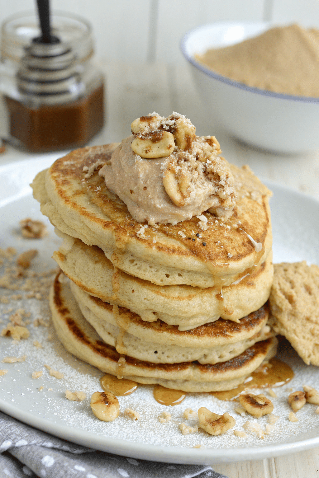 A photo of a plate of pancakes with a creamy topping, nuts, and a drizzle of syrup. The pancakes have a golden-brown hue and are stacked on a white plate. The toppings include a creamy mixture with nuts and a light dusting of powdered sugar. The plate is placed on a wooden surface, and in the background, there is a jar with a liquid substance and a bowl with a light brown substance. The style of the image is a close-up food photograph.