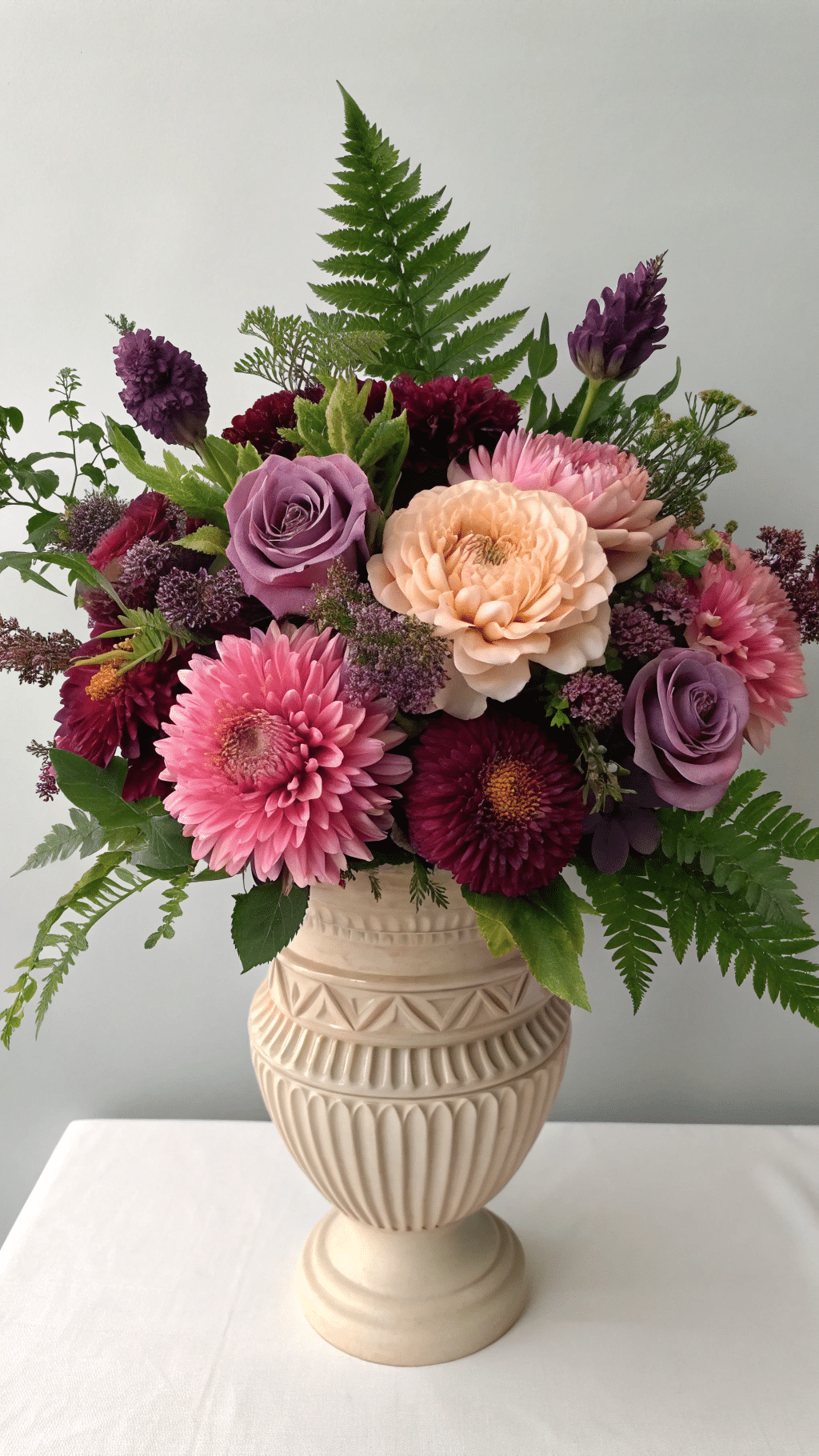 A vibrant dried flower arrangement featuring rich plums, mauve, and blush pinks, complemented by preserved ferns and greenery. The bouquet is large and lush, designed to be the focal point of a dining table.