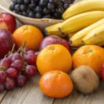 A photo of a variety of fresh fruits arranged on a wooden table. The fruits include apples with a mix of red and green hues, oranges with a vibrant citrus color, bananas with a bright yellow color, grapes with a deep purple color, and a pineapple with a unique, textured brown color. The arrangement is casual, showcasing the natural beauty and color palette of the fruits. The lighting is soft and natural, highlighting the textures and colors of the fruits.